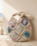A handmade crochet shoulder bag made of cream-colored granny squares with colorful floral patterns in blue, pink, and green, resting on a beige stone ledge.