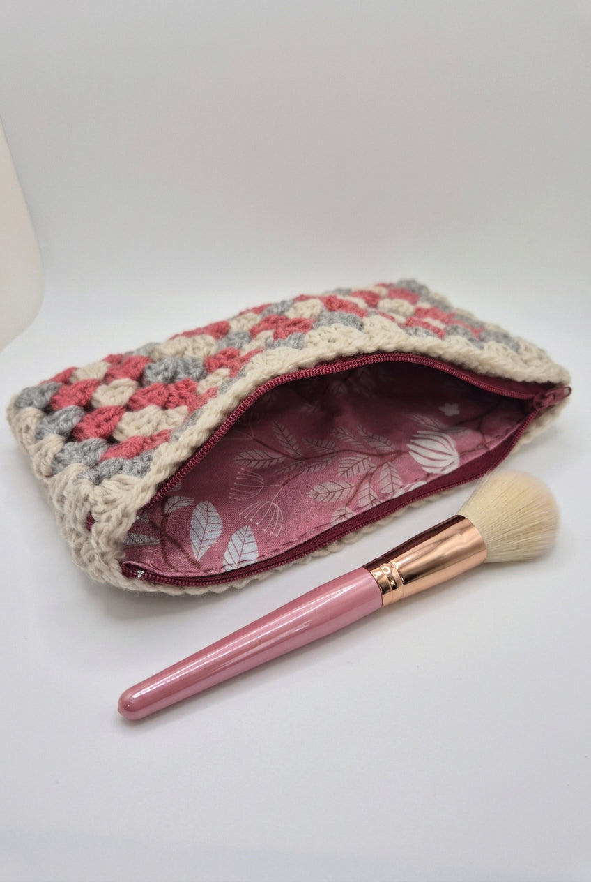 A handmade crochet pouch with a textured cream, pink, and gray exterior, featuring a burgundy zipper and a burgundy interior with a white leaf pattern.