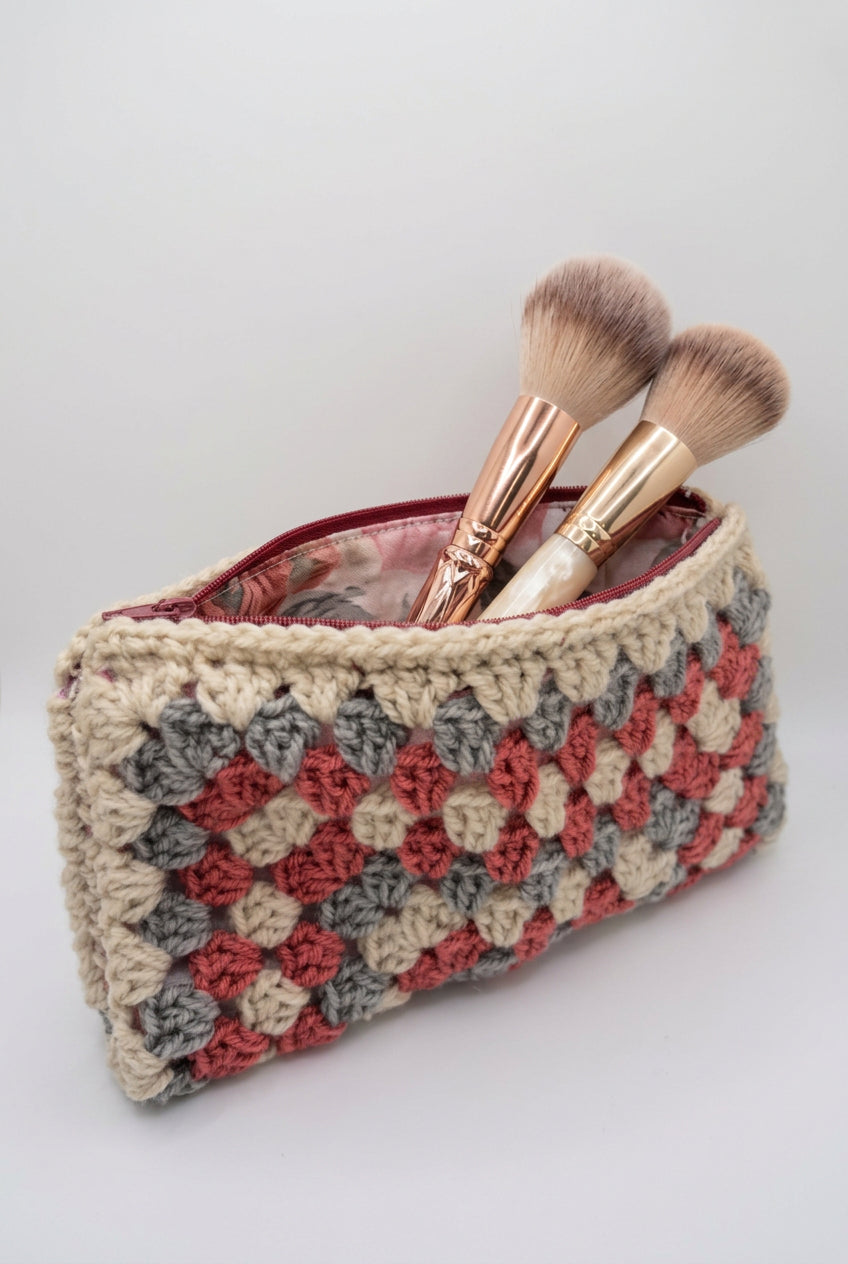 A handmade crocheted clutch bag with a textured checkerboard pattern in beige, gray, and red, featuring a solid red lining visible at the top, set against a plain light gray background.