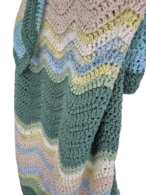 A close-up of a handmade crocheted blanket featuring a wavy chevron pattern in muted pastel shades of sage green, dusty blue, cream, and pale yellow.