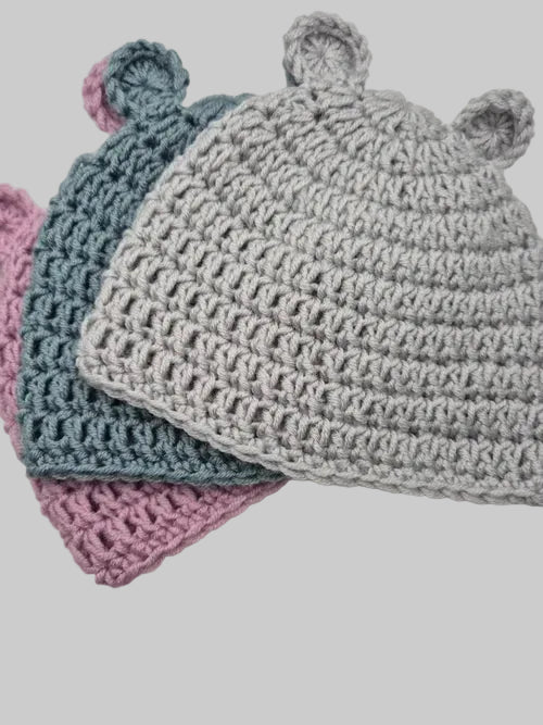 Three crocheted baby hats with ear details, shown overlapping on a light gray background. The hats are in light gray, dusty blue, and dusty pink.