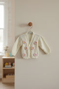 A cream-colored knit cardigan sweater for a baby or toddler, featuring pink crocheted flower appliqués with yellow centers, hanging on a wooden hanger against a softly blurred nursery background.