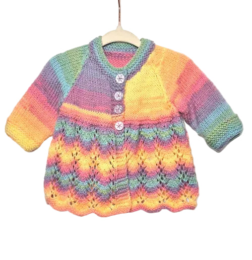 A handmade, long-sleeve baby cardigan sweater in a rainbow gradient yarn, hanging on a wooden hanger against a light gray fabric background. The cardigan features a V-neck, three decorative buttons, and a textured knit pattern with a scalloped hem.