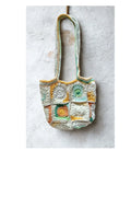 A handmade crocheted patchwork tote bag with long shoulder straps, featuring a mix of cream, green, yellow, and orange crochet squares, hanging against a textured white plaster wall.