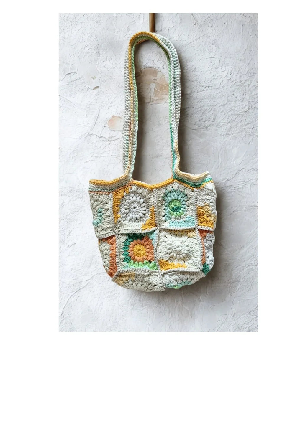 A handmade crocheted patchwork tote bag with long shoulder straps, featuring a mix of cream, green, yellow, and orange crochet squares, hanging against a textured white plaster wall.