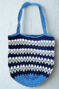A handmade crocheted tote bag with horizontal stripes in light blue, navy blue, and white, hanging against a textured white wall.