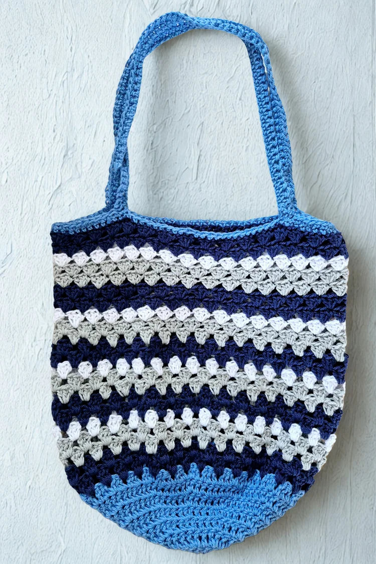 A handmade crocheted tote bag with horizontal stripes in light blue, navy blue, and white, hanging against a textured white wall.